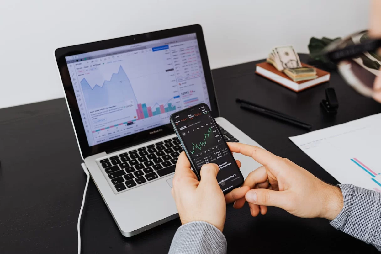Person holding a smartphone in front of laptop, both displaying financial charts