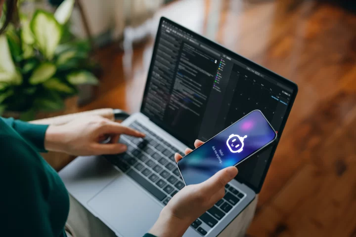 Asian woman using AI chatbot virtual assistance app on smartphone to assist her while working on laptop. Using AI technology while working at home. Online robot system. Concept of VR and AR technologies. People, AI, innovation and technology